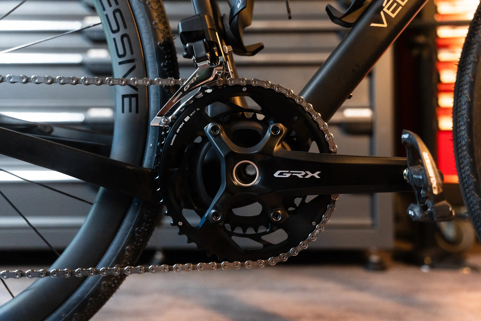 Close-up of a black GRX bicycle crankset with a chainring and chain, attached to Vélobsessive's Gravel Bike  frame in a workshop setting.