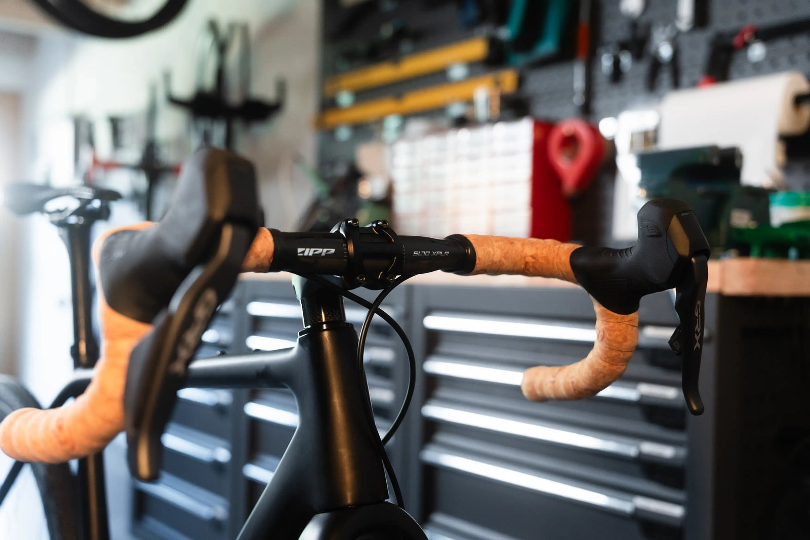 Close-up of Vélobsessives Gravel Bike Cobblestone drop handlebar with black components and tan grip tape, set in a workshop with blurred tool cabinets and wall-mounted equipment in the background.