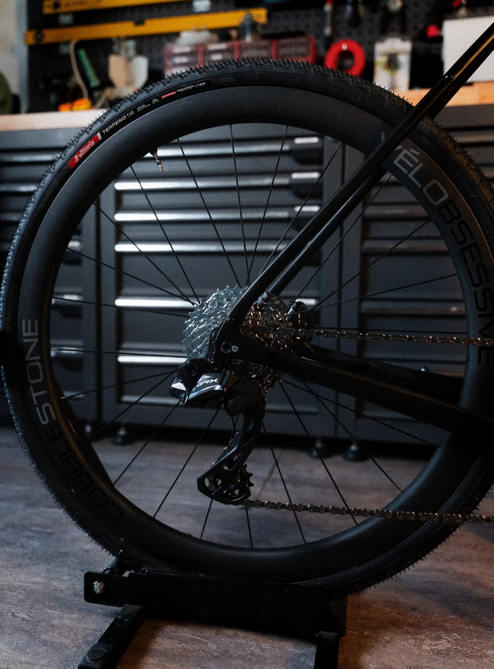 Close-up of Vélobsessive's Gravel Bike Cobblestone wheel and rear hub mounted on a stand in a workshop, with tool cabinets in the background. 