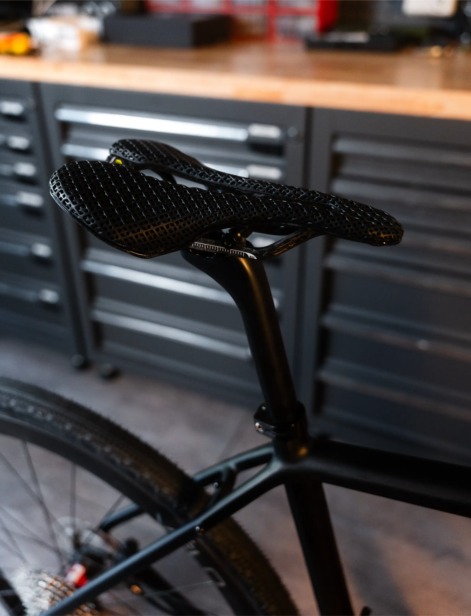 Close-up of Vélobsessive's Gravel Bike  saddle and seatpost on a matte black bicycle frame, photographed in a workshop with dark tool cabinets in the background.