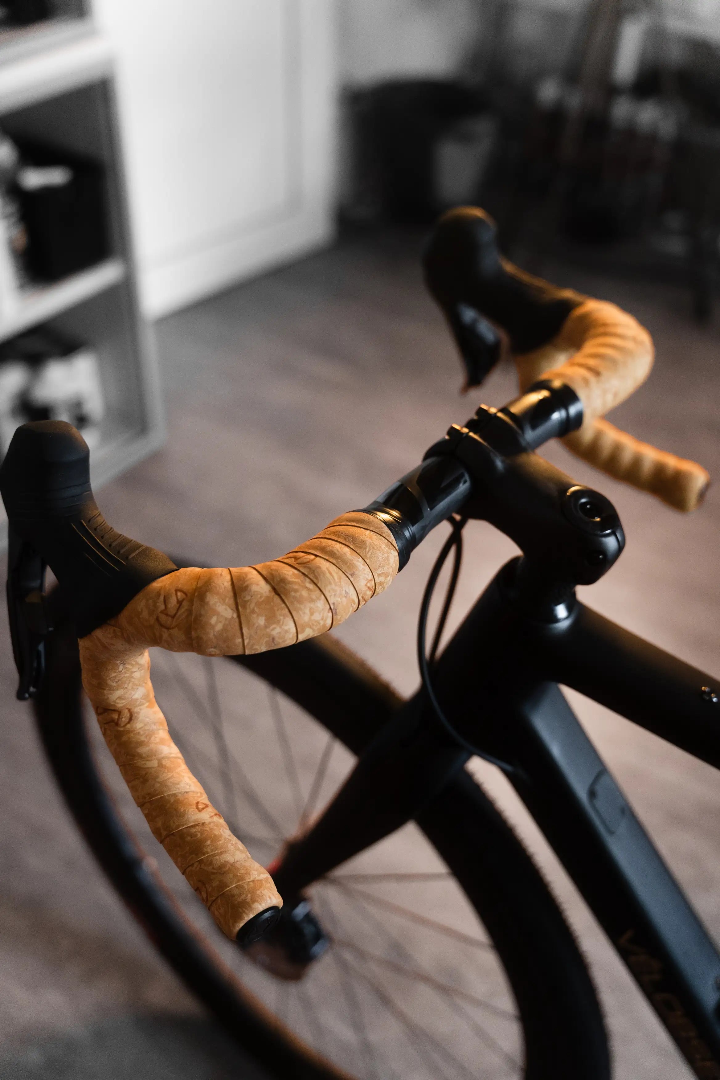 Close-up of Vélobsessive's Gravel Bike  handlebar area with tan cork-wrapped drop handlebars and black brake levers, photographed indoors with a blurred room background.