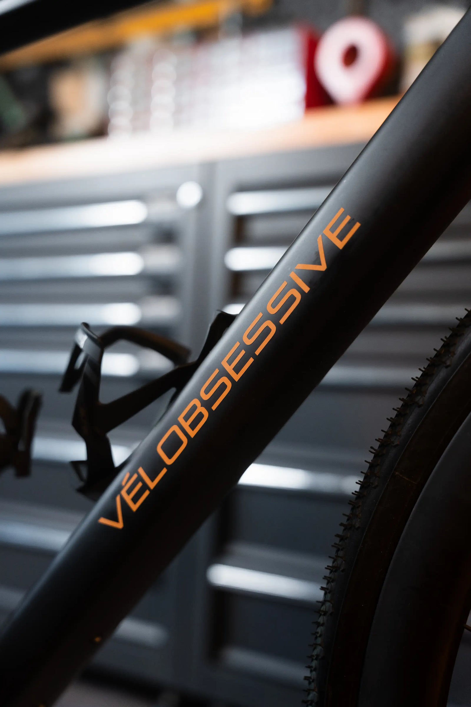 Close-up of Vélobsessives Gravel Bike Cobblestone frame with the word 'VELOOBSESIVE' printed in orange, with a blurred workshop or garage background.